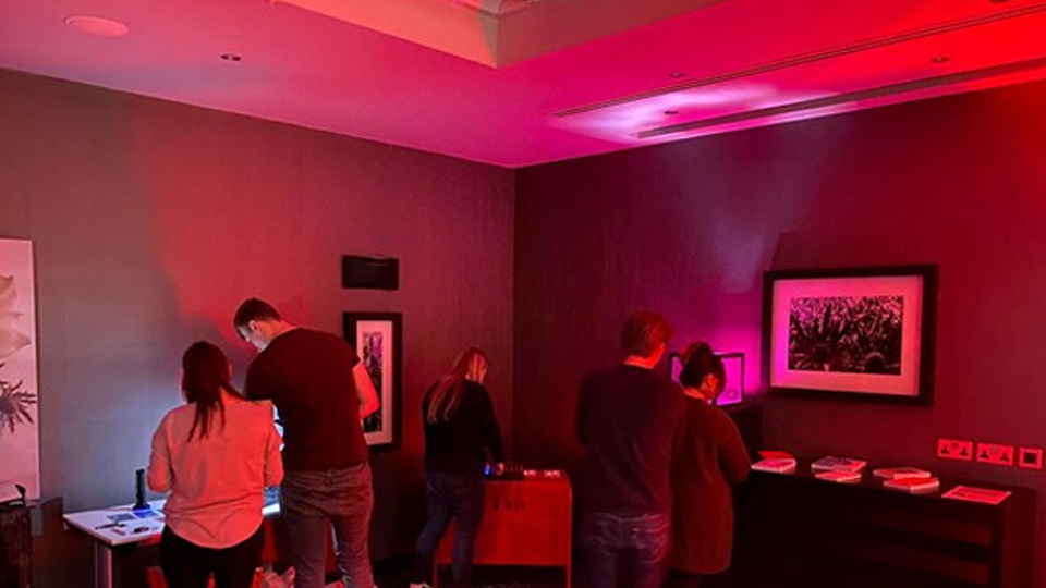 Several people in a room looking at items on tables. The room is lit red. 